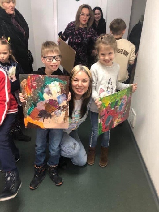 Children displaying colorful paintings with a smiling adult in a classroom setting.