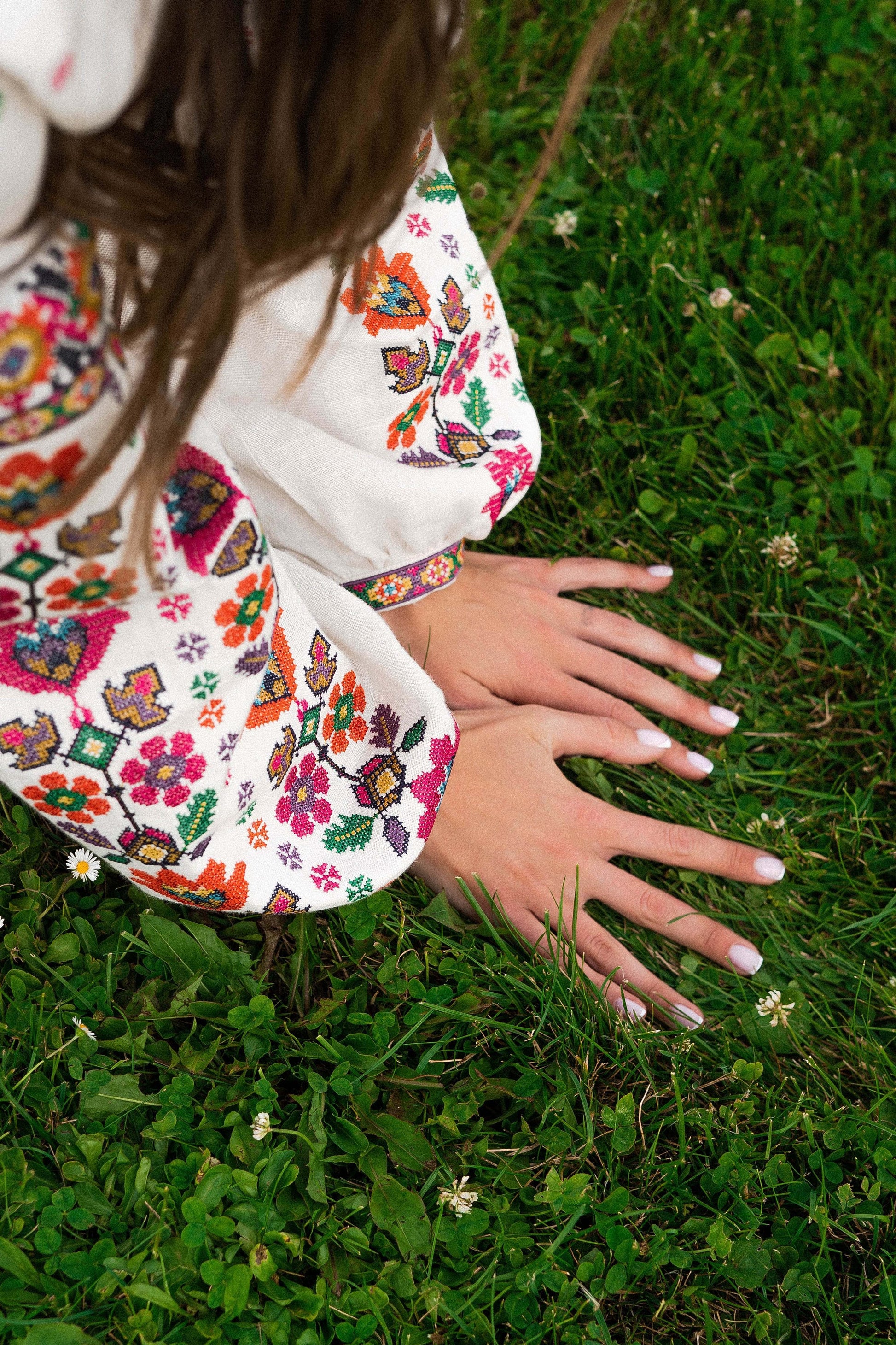 Woman wearing embroidered blouse with colorful floral patterns, hands resting on green grass.