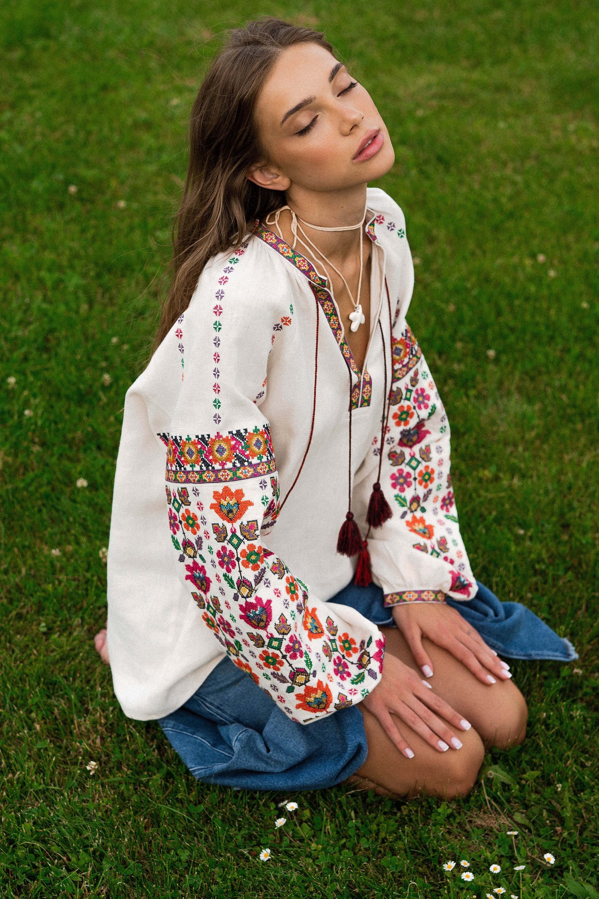 Woman wearing a soft beige linen Vyshyvanka blouse with colorful Ukrainian embroidery, sitting on grass in summer.