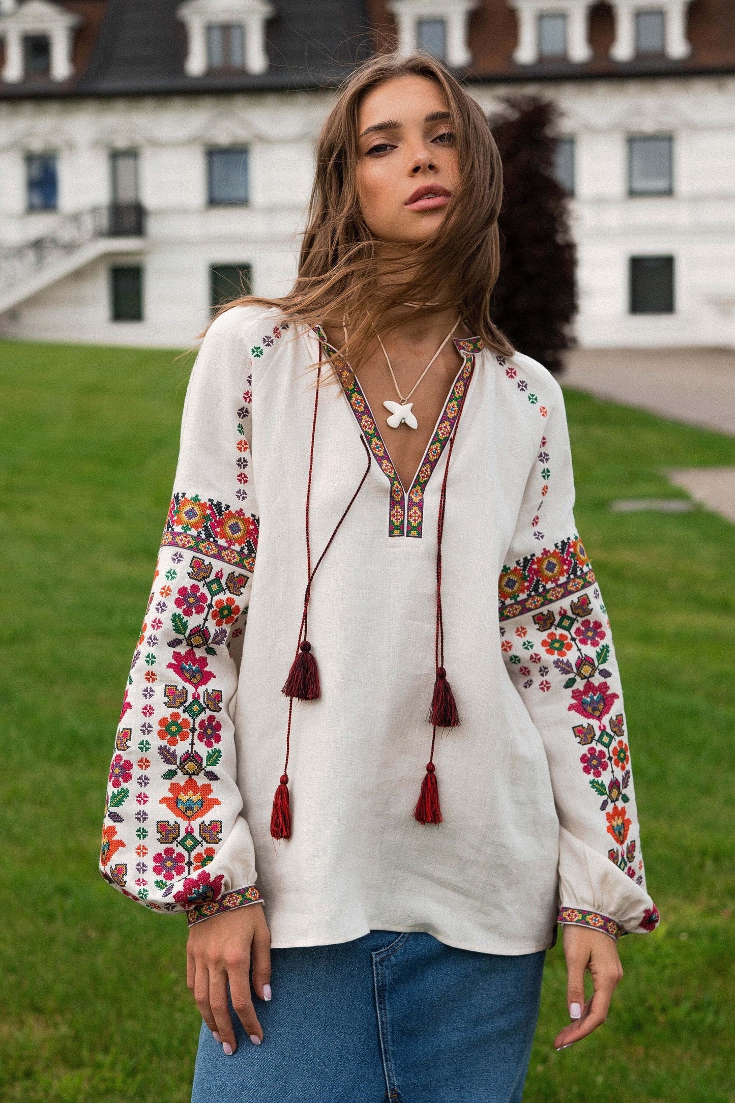 Woman wearing beige linen Vyshyvanka blouse with colorful embroidery, standing outdoors near a historic building.