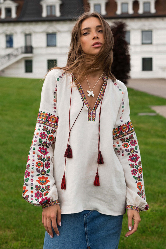 Woman wearing beige linen Vyshyvanka blouse with colorful embroidery, standing outdoors near a historic building.