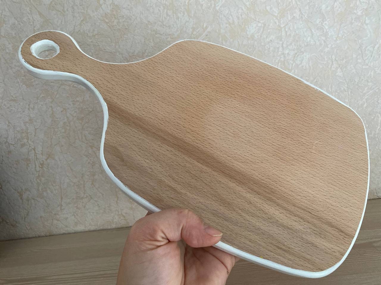 Hand holding a blank wooden cutting board with a handle against a beige background, ready for decorative painting.