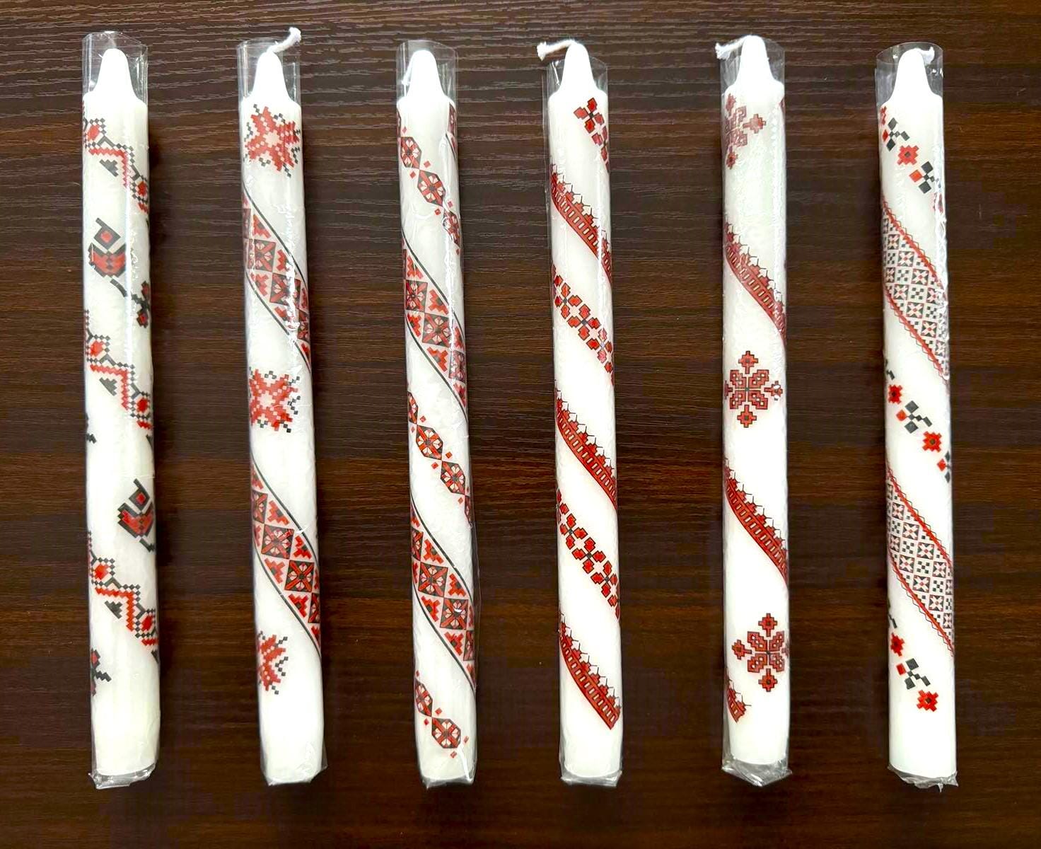 Handmade Ukrainian holiday taper candles with traditional red and white festive designs displayed on a wooden surface.