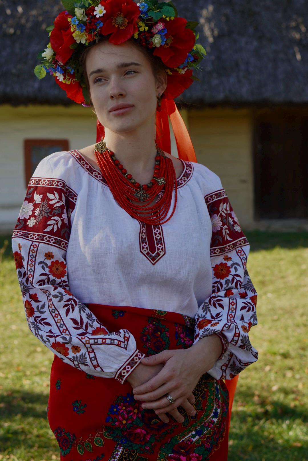 Woman in traditional Ukrainian attire with a handmade red multi-row necklace and flowered headdress, standing outdoors.