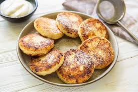 Plate of golden Ukrainian syrnyky cheese pancakes, lightly dusted with powdered sugar, served with a dollop of sour cream.