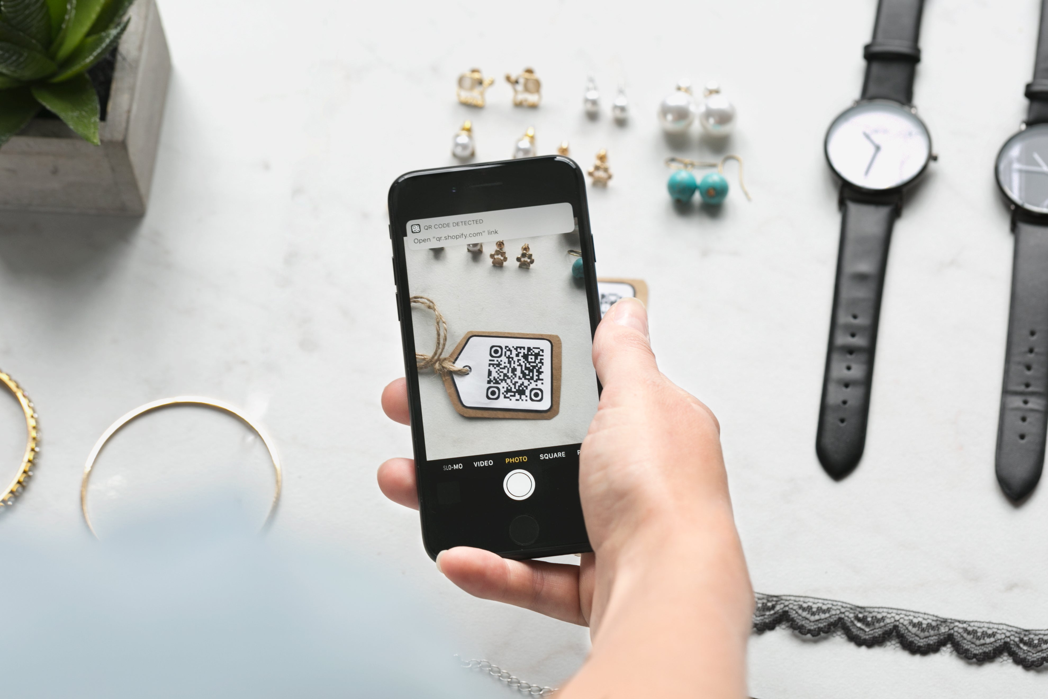 Person scanning QR code on smartphone surrounded by jewelry and watches on a marble surface.