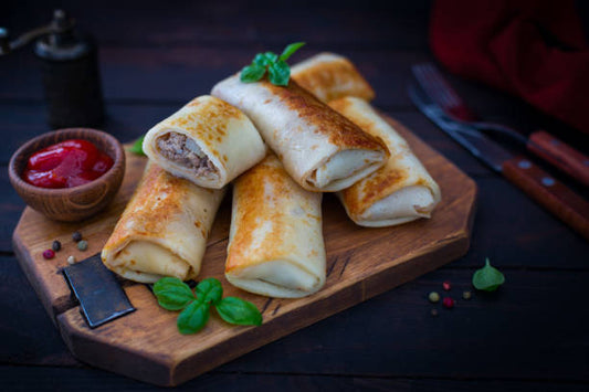 Homemade Ukrainian crepes with beef and chicken fillings on a wooden board, garnished with herbs and served with a side of ketchup.