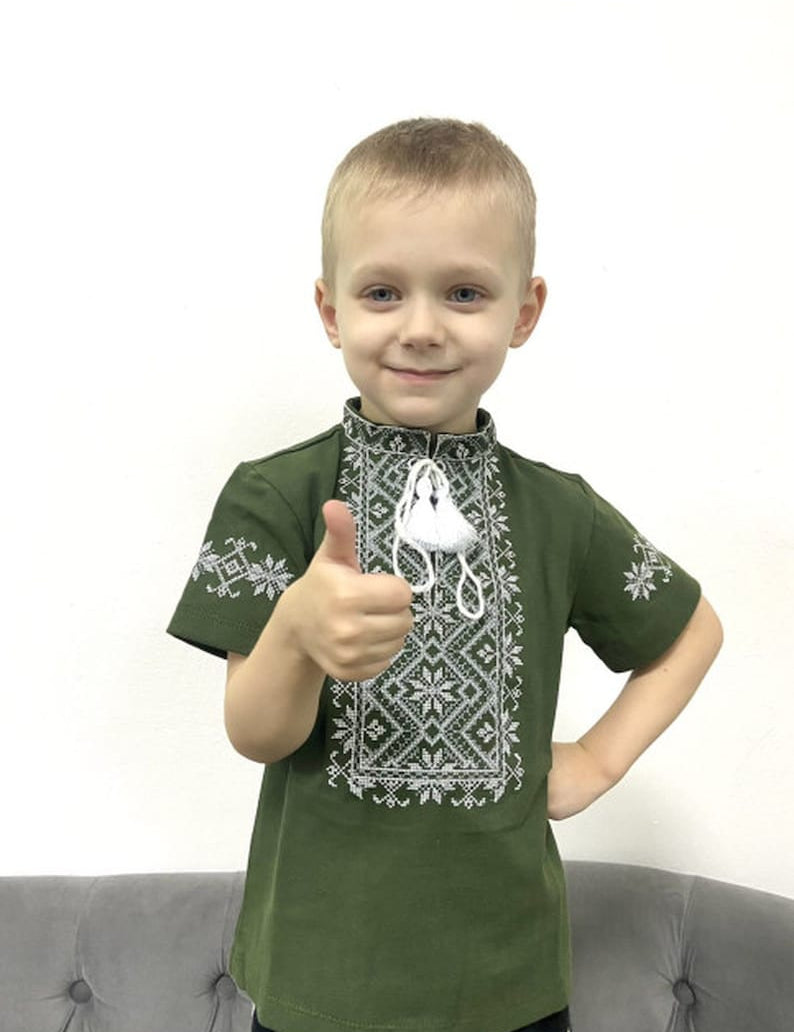 Young boy wearing a khaki Ukrainian embroidered shirt with short sleeves and traditional cross-stitch patterns, giving a thumbs up.