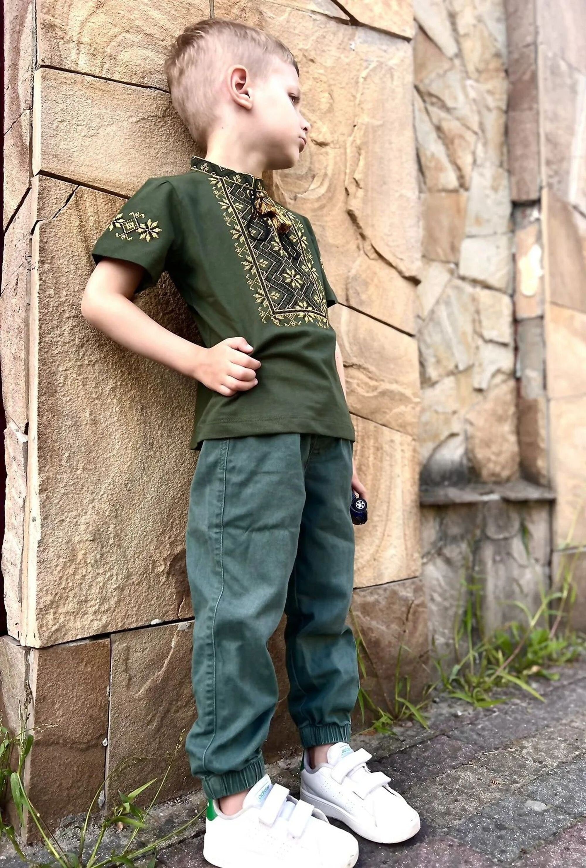 Boy wearing a khaki Ukrainian embroidered Vyshyvanka shirt with geometric patterns, short sleeves, and stand-up collar.