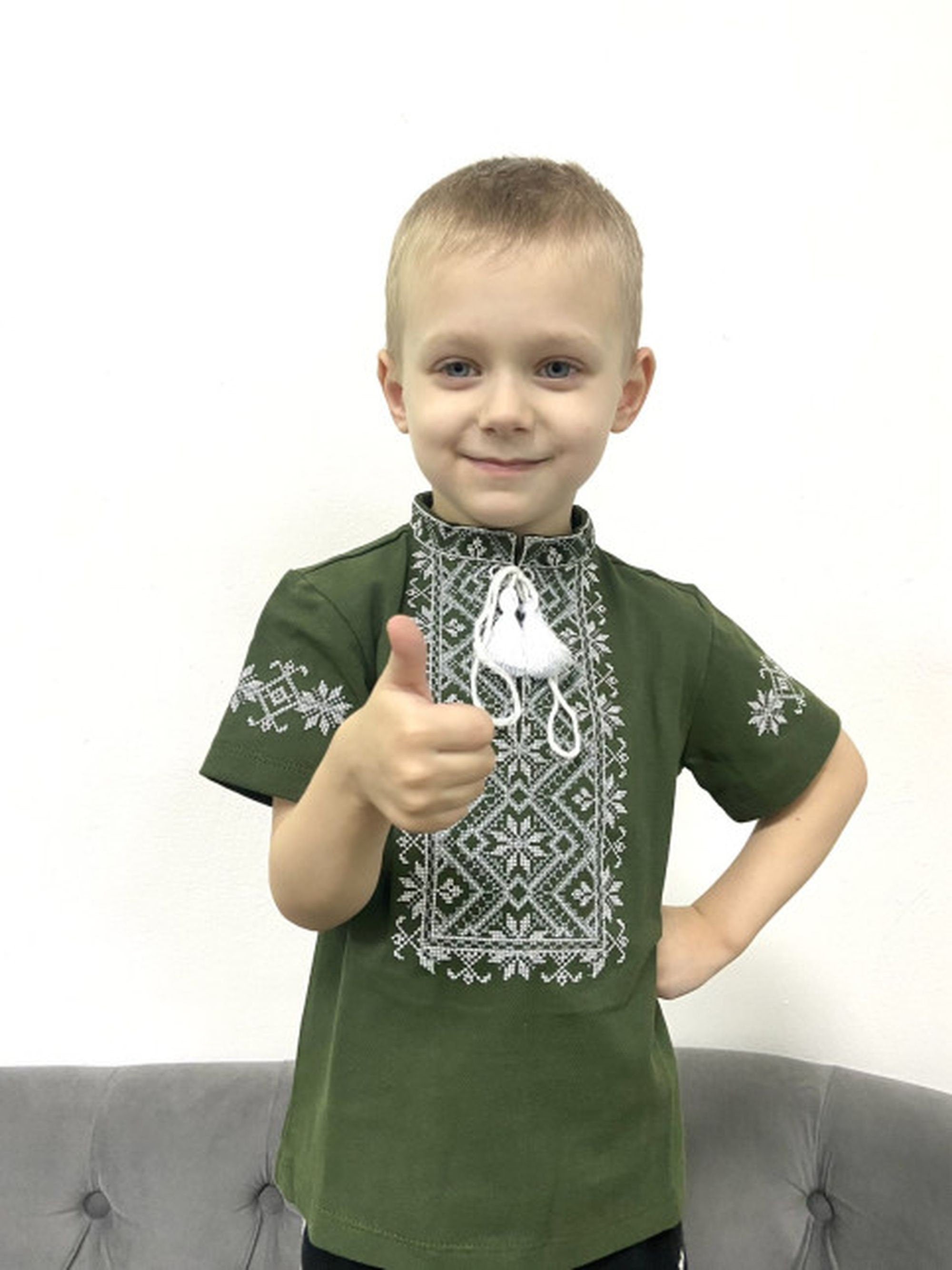 Boy wearing a khaki Ukrainian embroidered shirt with geometric cross-stitch design, short sleeves, and stand-up collar, giving a thumbs-up.