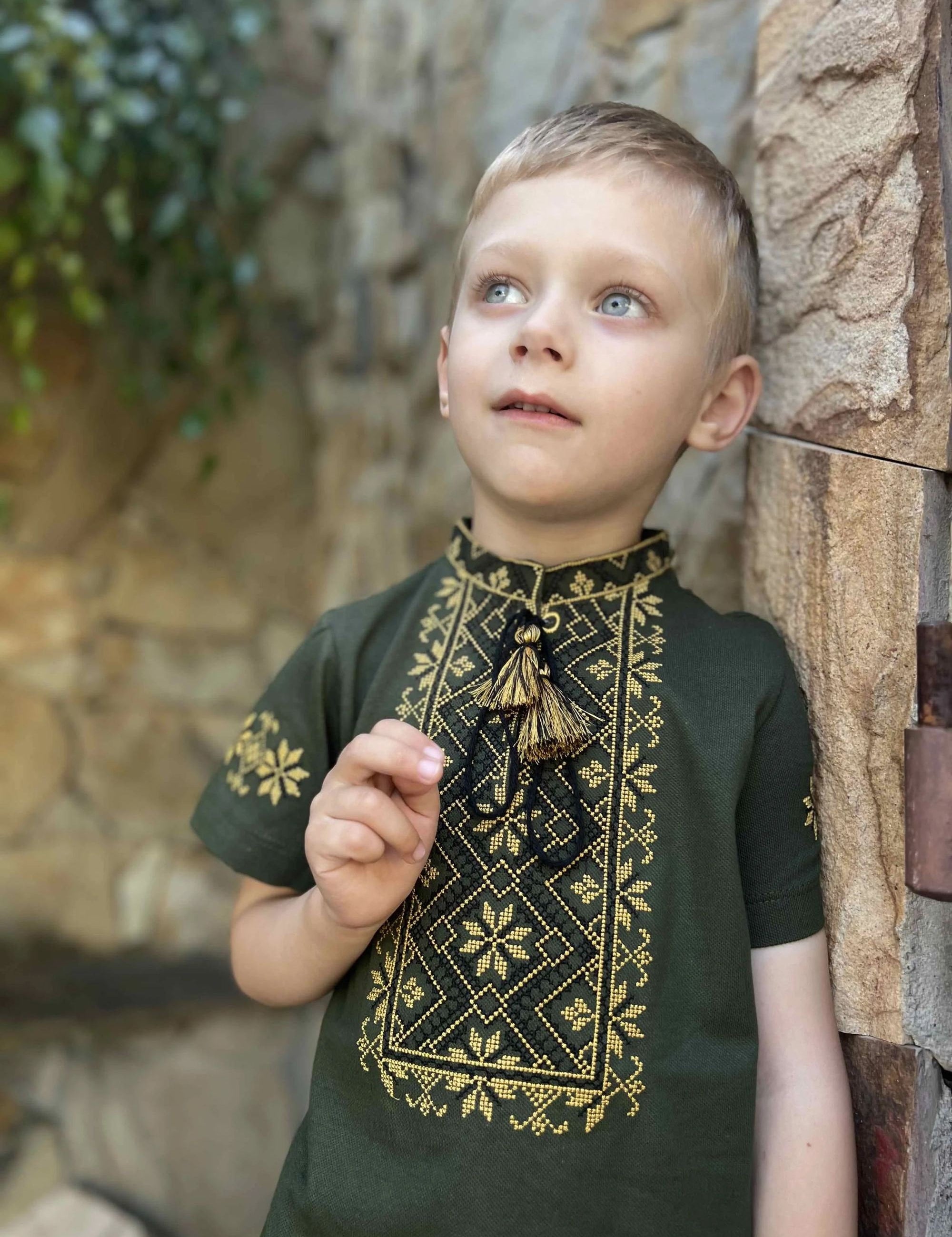 Boy wearing a khaki Ukrainian embroidered shirt with short sleeves and geometric cross-stitch design, standing outdoors.