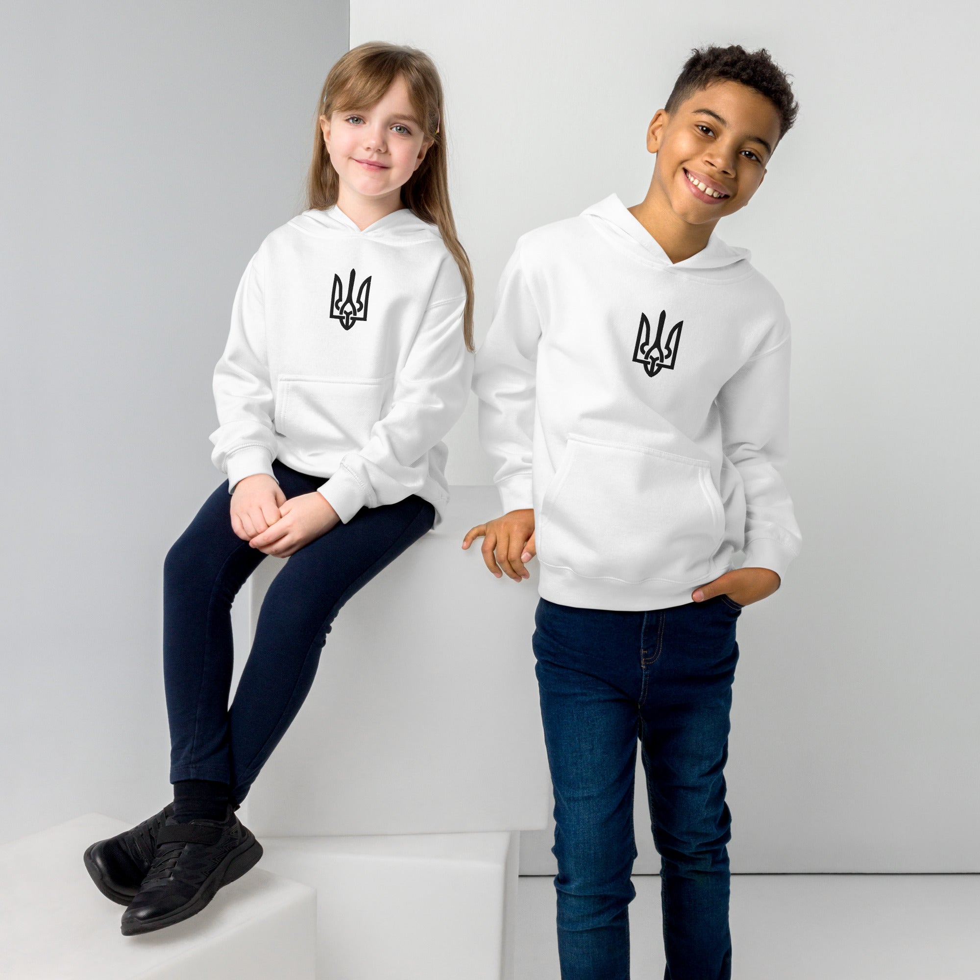 Kids wearing white fleece hoodies with Ukrainian Tryzub emblem, smiling and standing on a white platform.