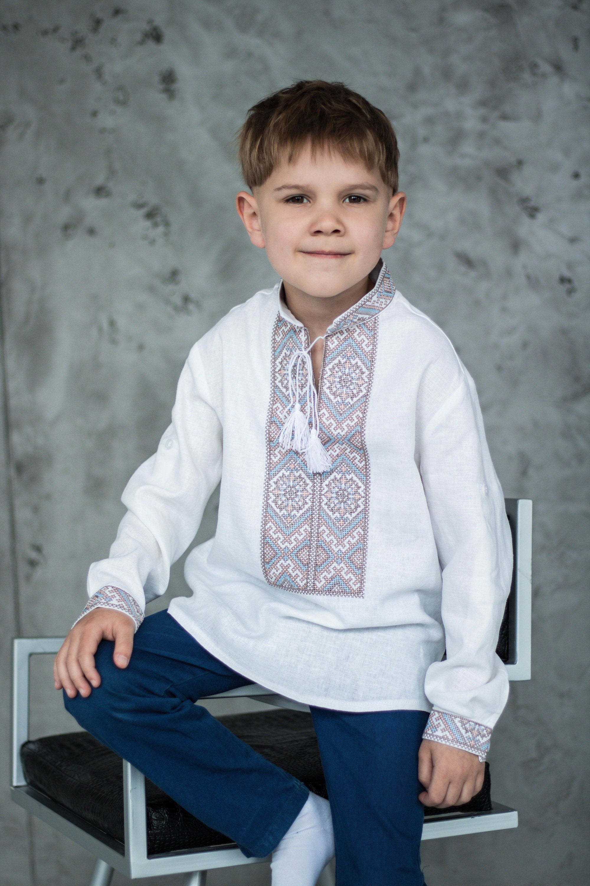 Young boy wearing a traditional Ukrainian Vyshyvanka shirt with blue and brown geometric embroidery, seated on a chair.