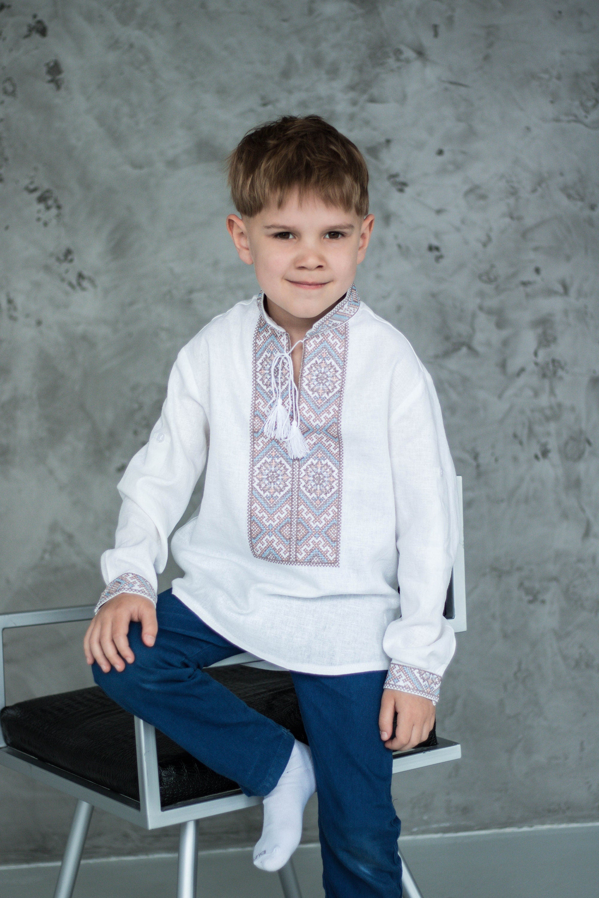 Young boy wearing a Ukrainian Vyshyvanka shirt with blue and brown embroidery, sitting on a chair.