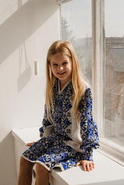 Girl wearing a traditional Ukrainian embroidered knitted dress, sitting by a window, showcasing blue and white patterns.