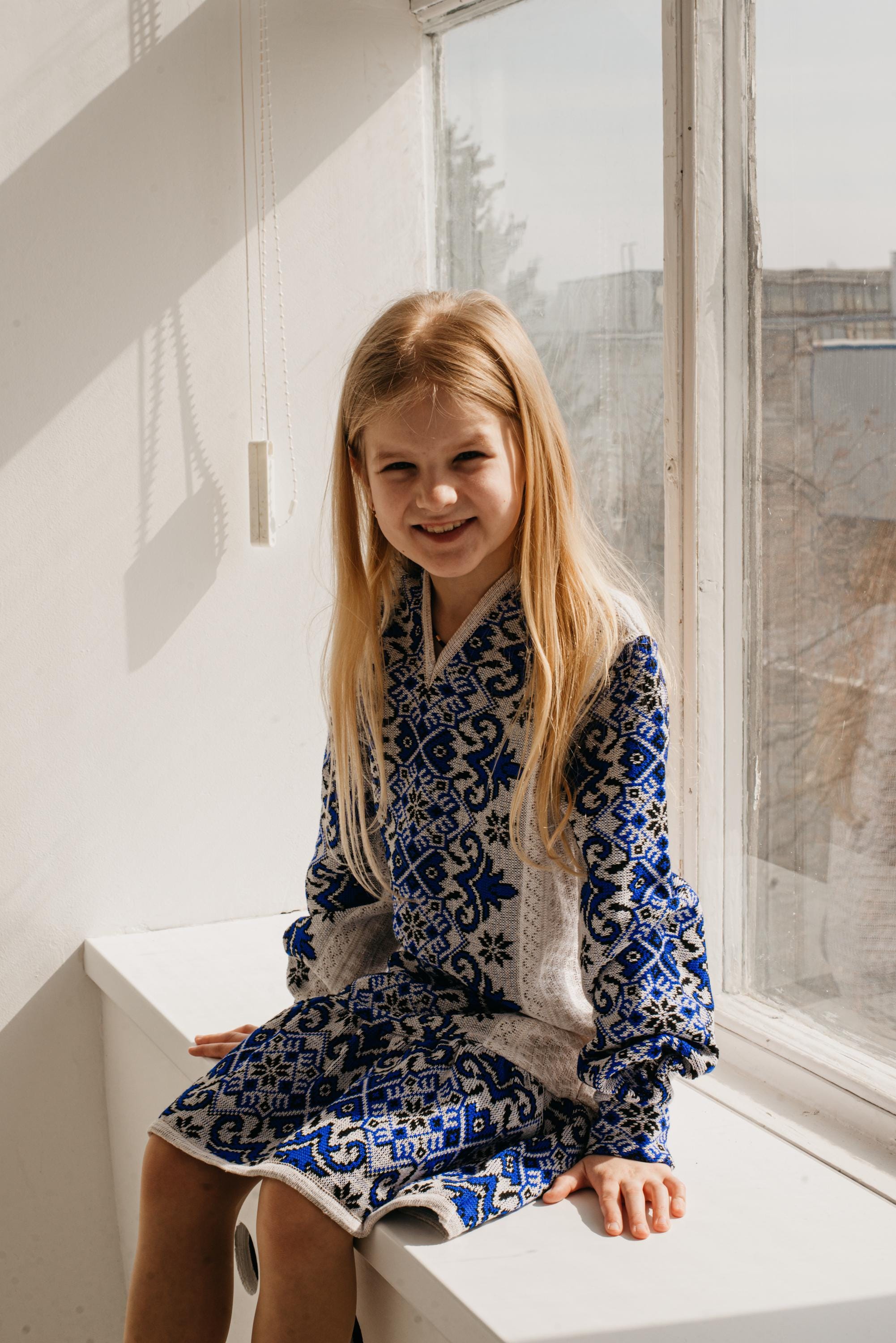 Girl wearing a traditional Ukrainian embroidered knitted dress, sitting by a window, showcasing blue and white patterns.