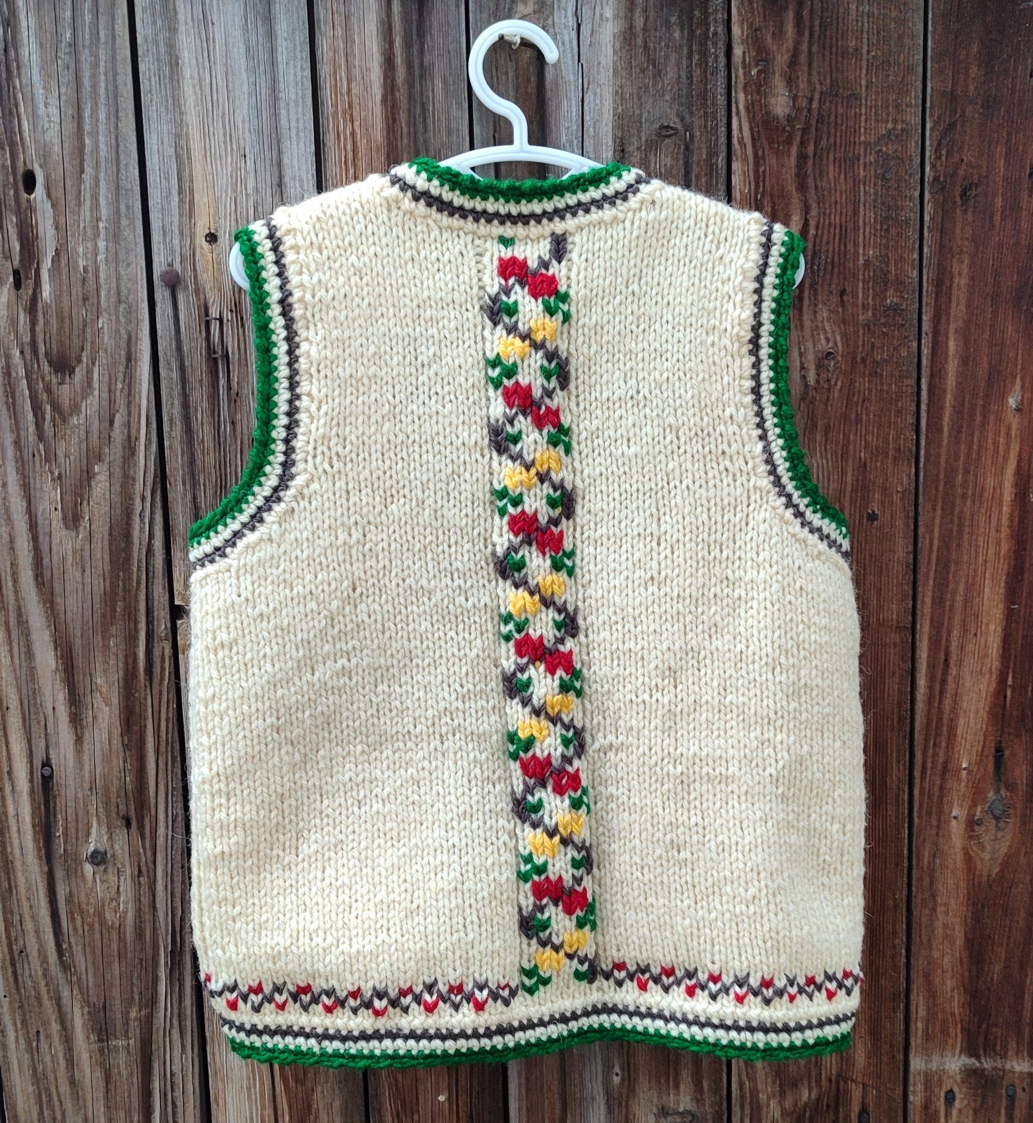 Hand-knitted unisex folk vest for kids, size 7-8, made from 100% wool with colorful embroidery and green crochet trim, hanging on a wooden background.