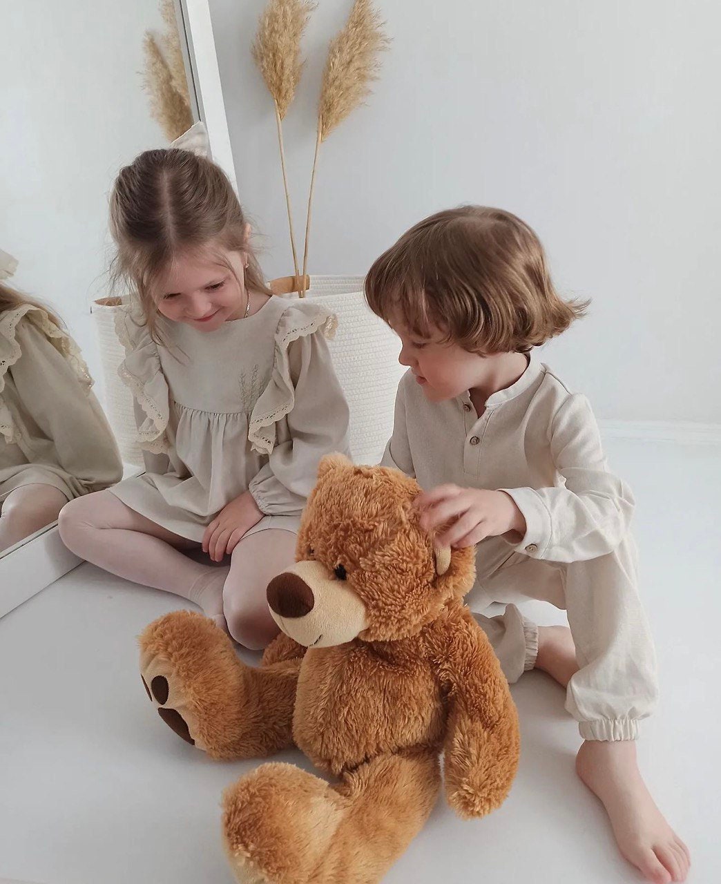 Two children in neutral-toned outfits sitting on the floor with a teddy bear, reflecting cozy and comfortable kids' fashion in a minimalist room.