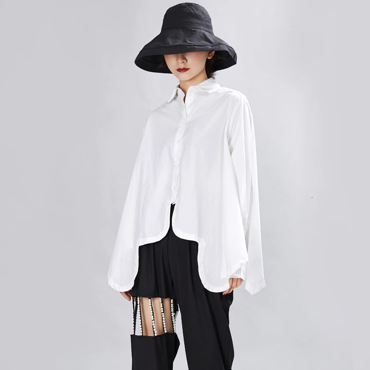 Woman wearing Valcira Irregular Hem Shirt in white with a flowy design, paired with black pants and a wide-brimmed hat. Fashionable outfit.
