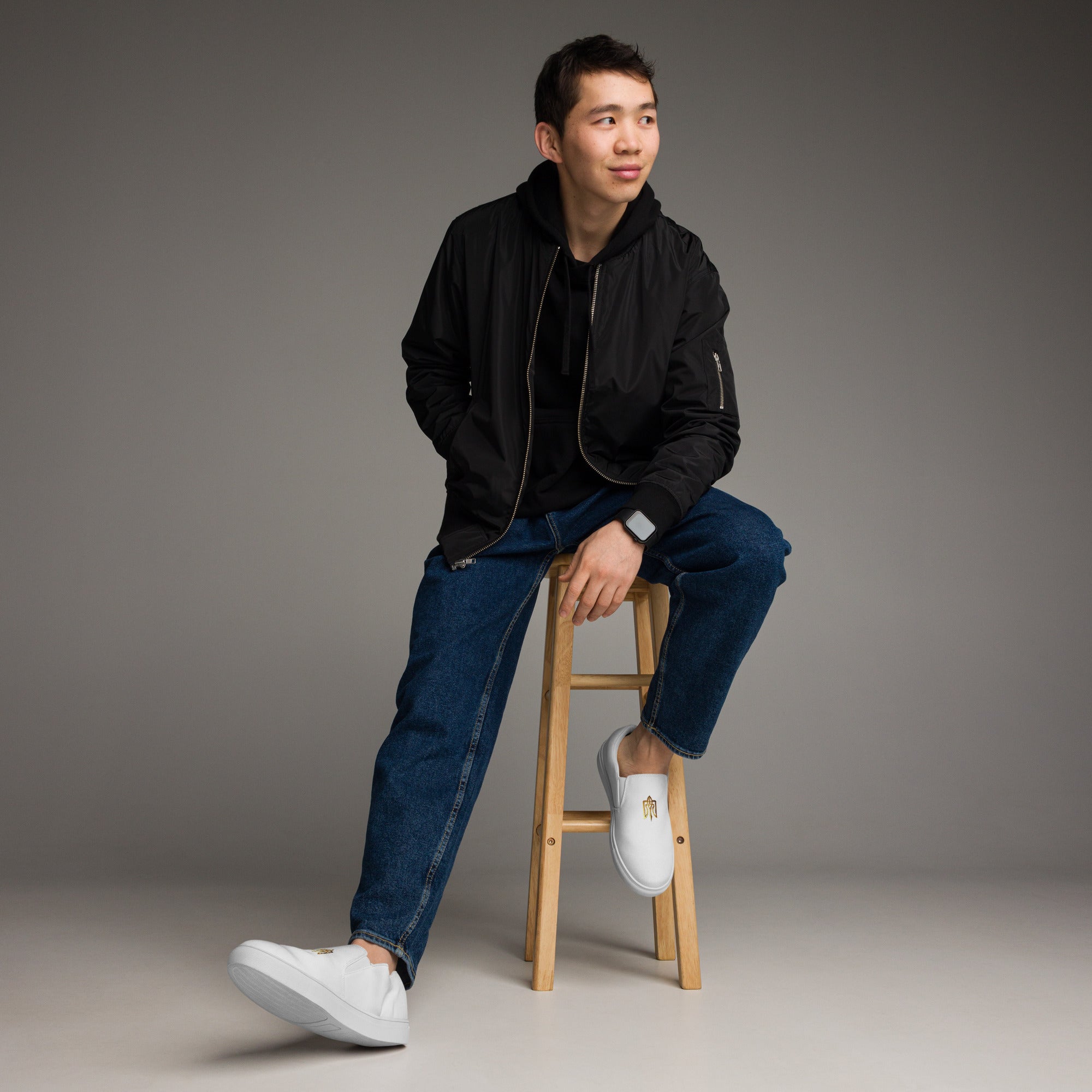 Man wearing Gold Ukrainian Trizub Slip-On Canvas Shoes sitting on a wooden stool against a gray background.