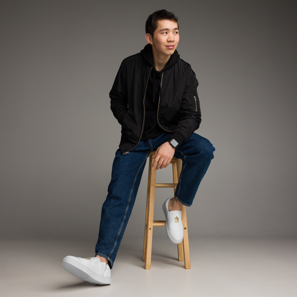 Young man sitting on stool wearing Gold Ukrainian Trizub slip-on canvas shoes with a black jacket and jeans.