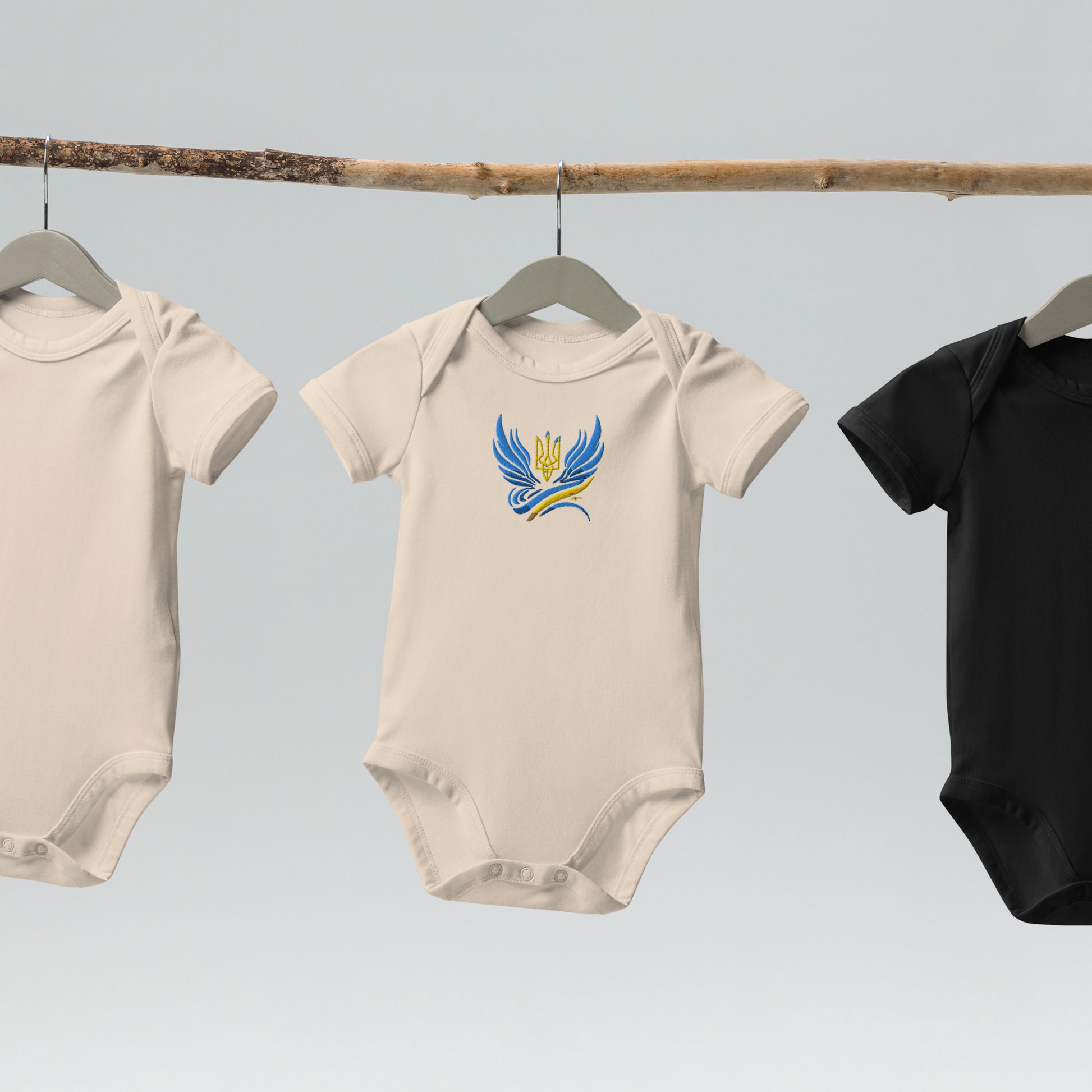 Organic cotton baby bodysuits with Ukrainian Tryzub logo in cream and black hanging on a natural wooden rod.