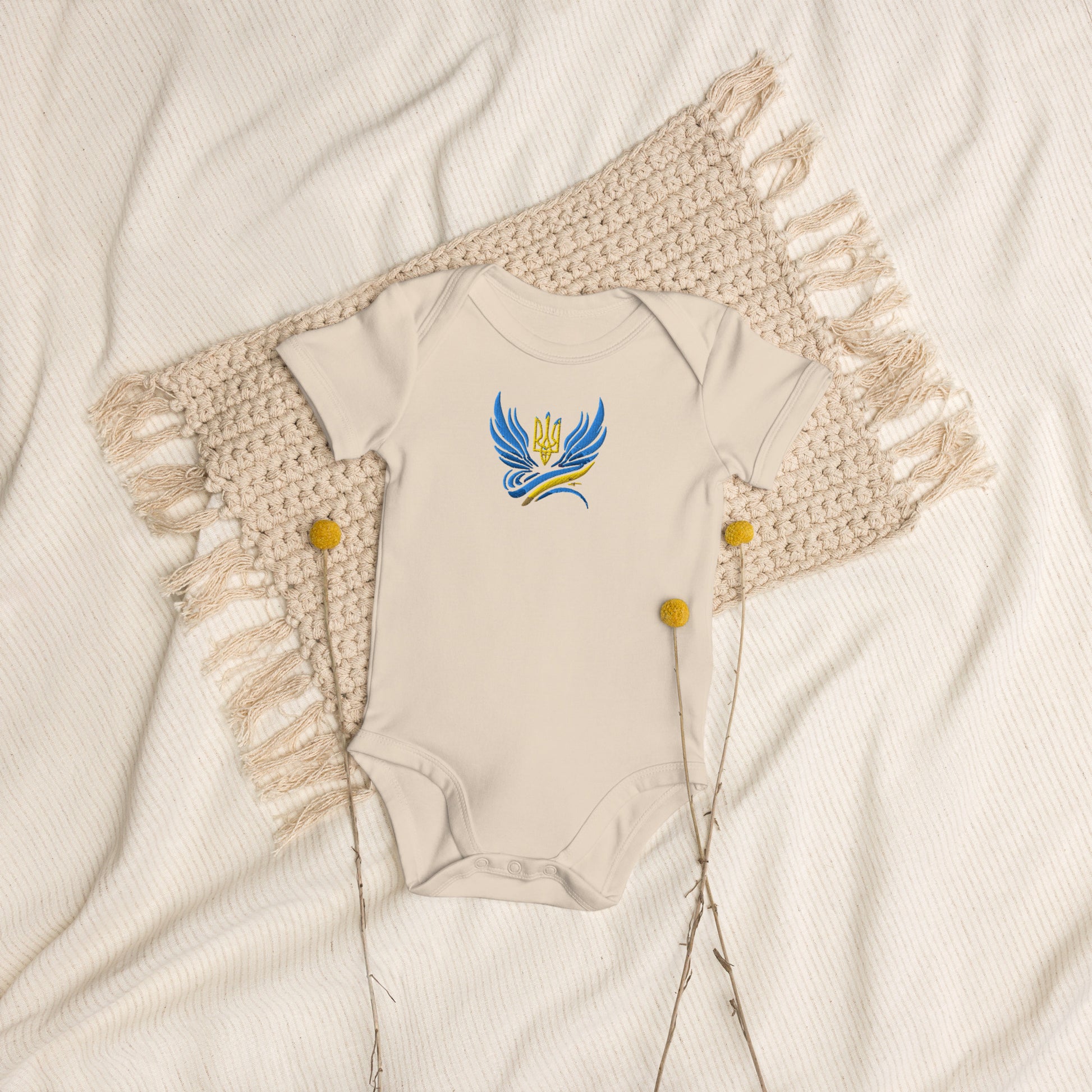 Organic cotton baby bodysuit with Ukrainian Tryzub logo on beige blanket, showcasing sustainable and stylish Ukrainian heritage attire.