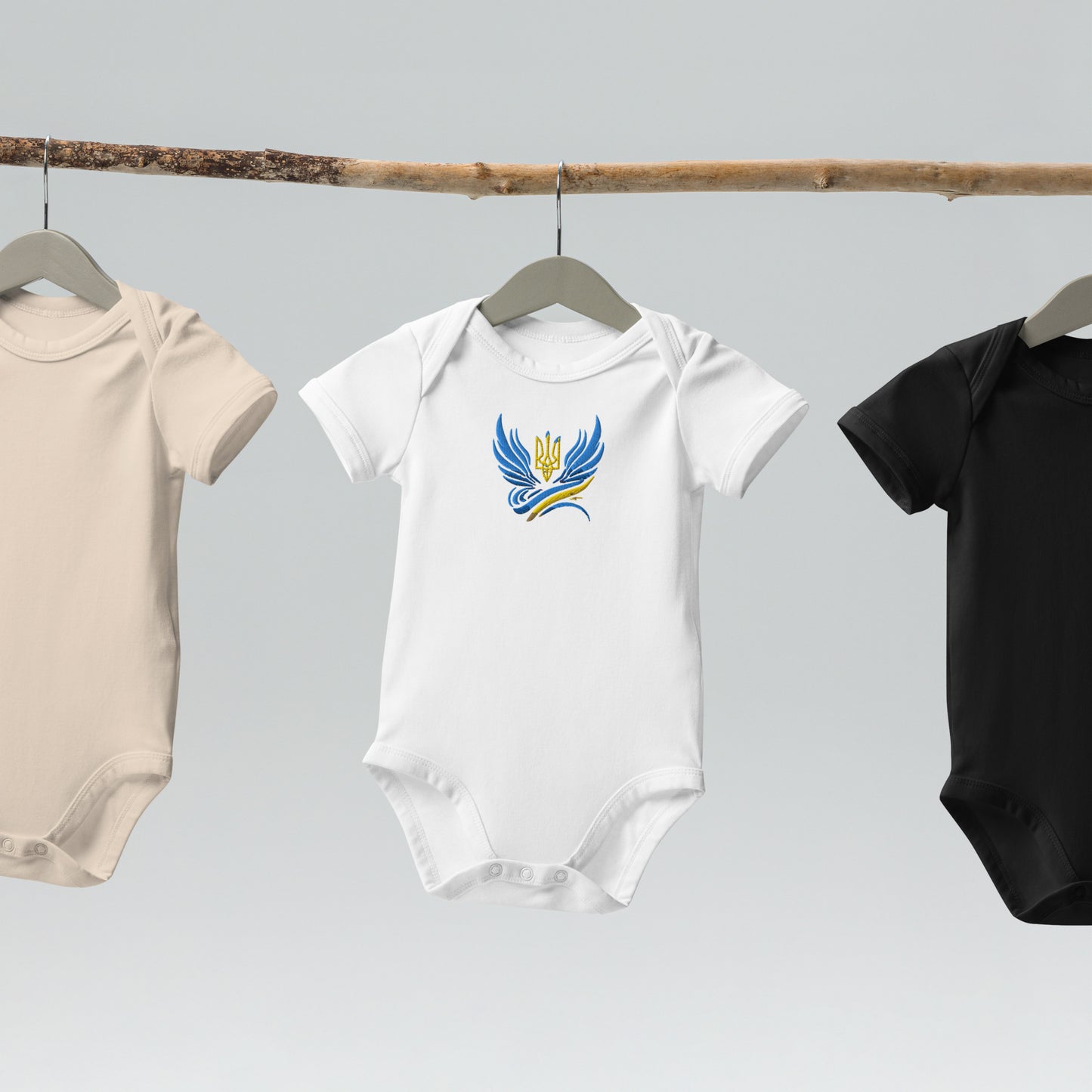 Organic cotton baby bodysuits hanging on a rod, featuring Ukrainian Tryzub logo in white, beige, and black colors.