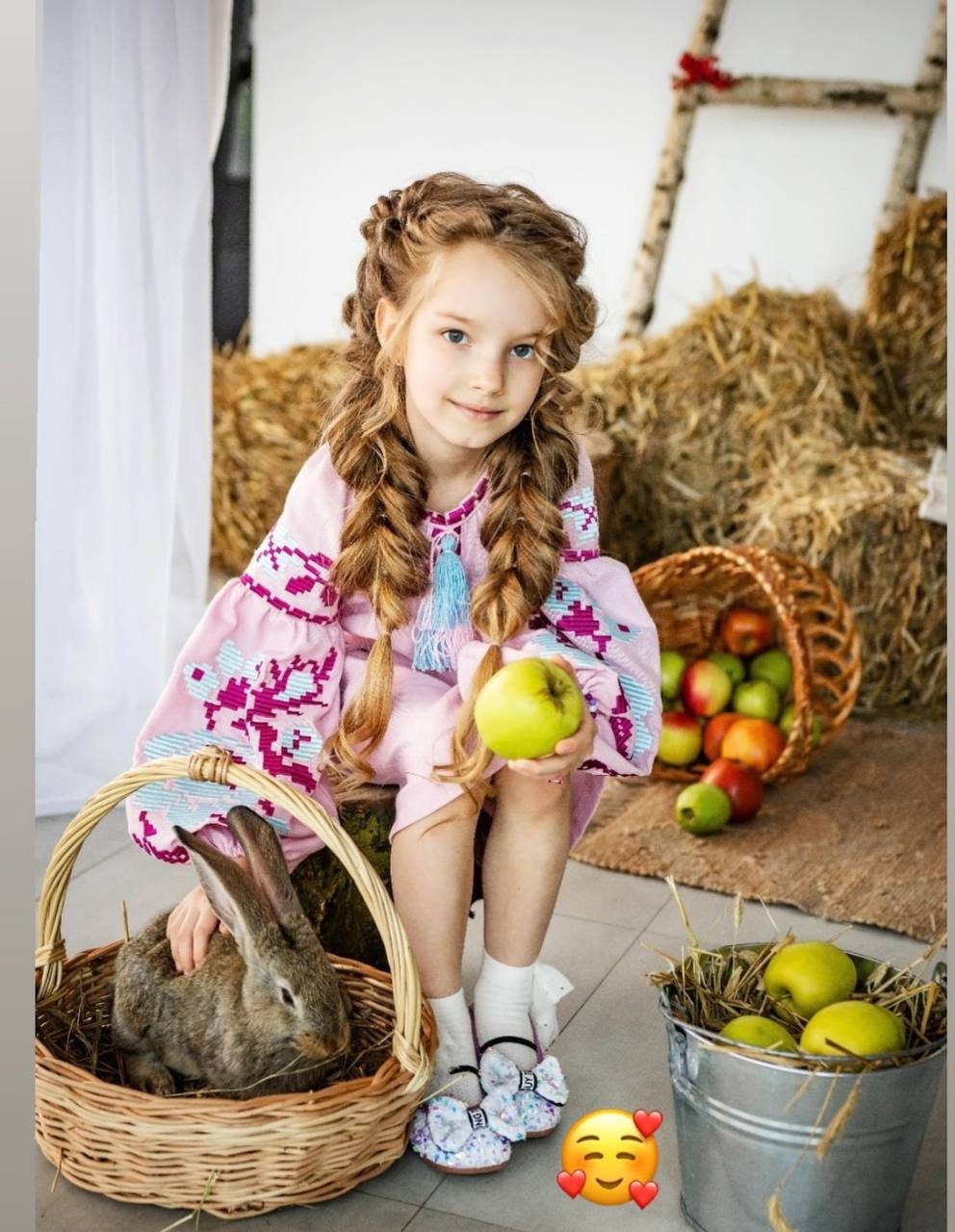 Girl in pink embroidered dress holding an apple next to a rabbit in a basket with hay and apples.