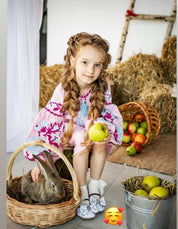 Girl in pink embroidered dress holding an apple next to a rabbit in a basket with hay and apples.