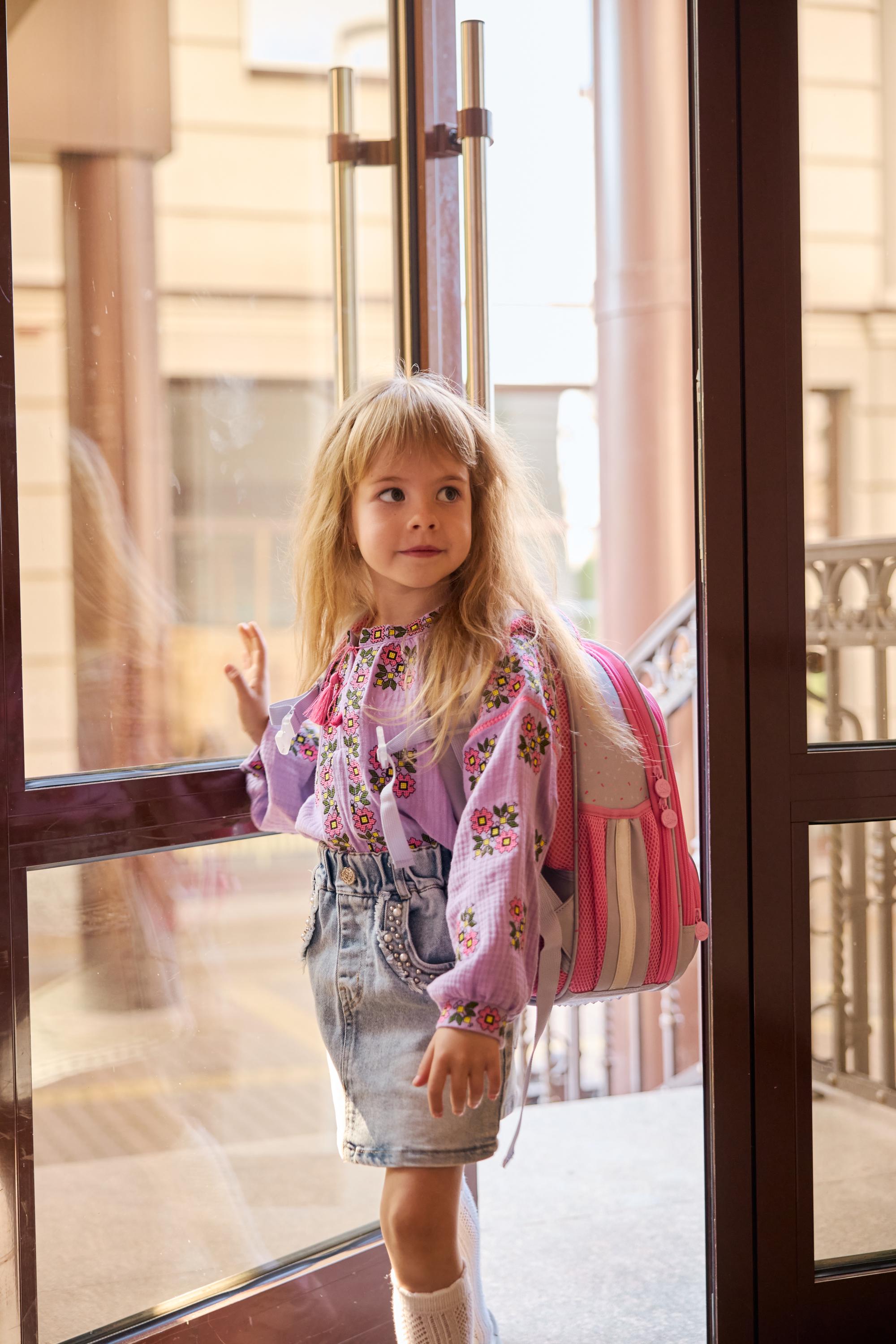 Girl wearing a lavender embroidered muslin vyshyvanka blouse with floral design, standing at a door with a pink backpack.