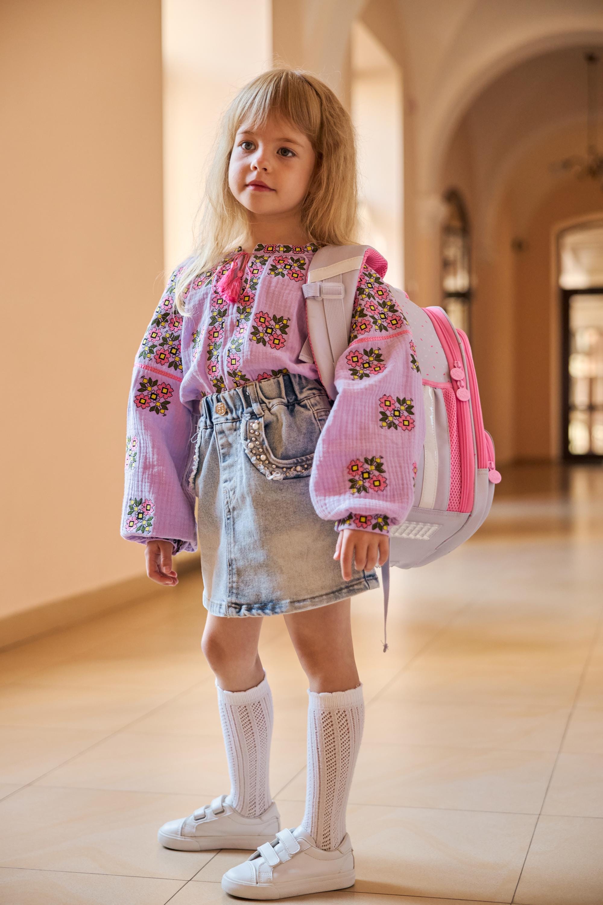 Girl wearing lavender embroidered muslin vyshyvanka blouse, denim skirt, white socks, and sneakers, standing indoors with a pink backpack.