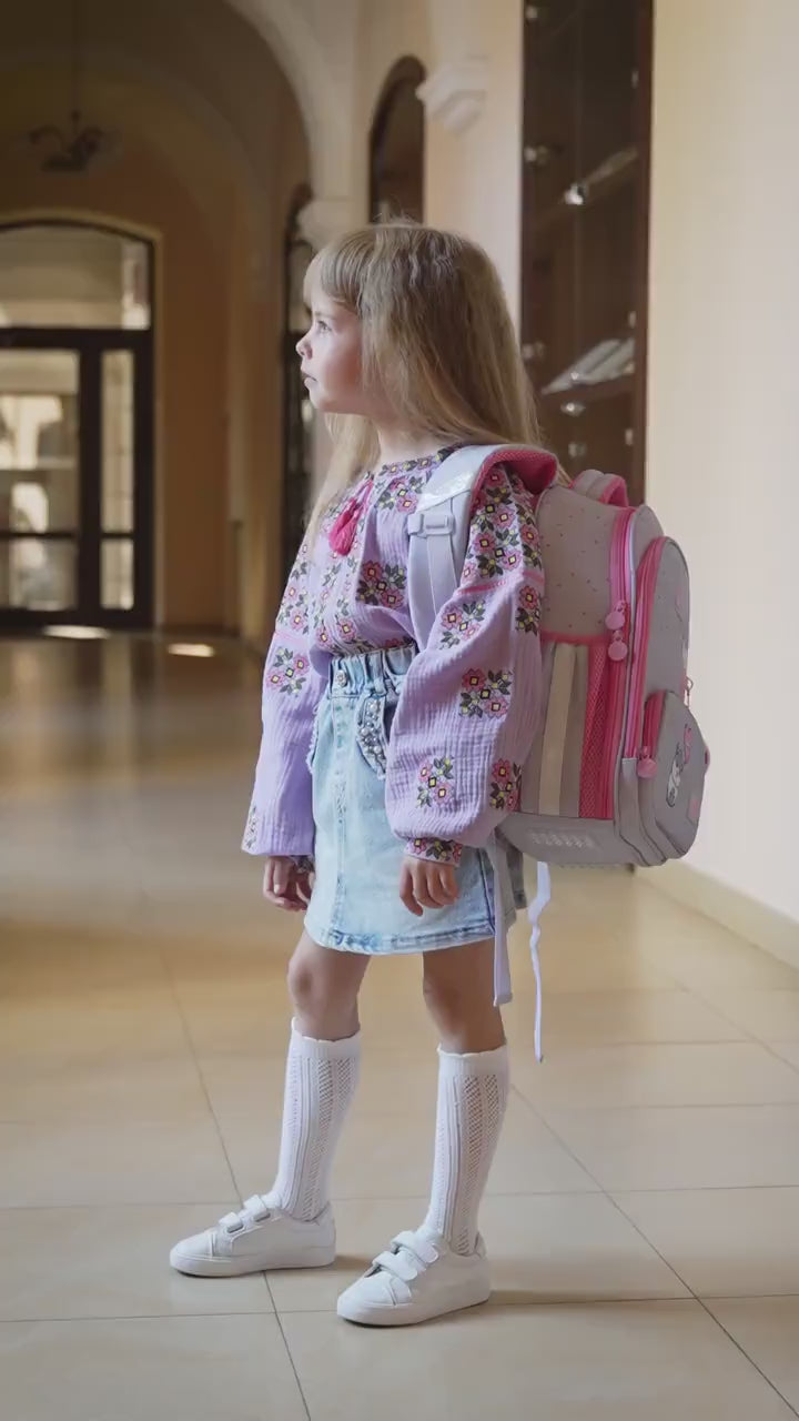 Girl wearing lavender embroidered vyshyvanka blouse with floral designs and denim skirt, carrying a pink backpack in a hallway.