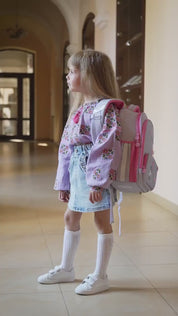 Girl wearing lavender embroidered vyshyvanka blouse with floral designs and denim skirt, carrying a pink backpack in a hallway.