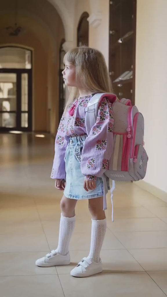 Girl wearing lavender embroidered vyshyvanka blouse with floral designs and denim skirt, carrying a pink backpack in a hallway.
