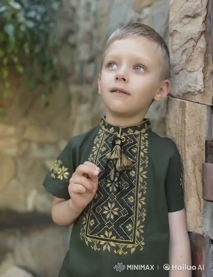 Boy wearing khaki Ukrainian embroidered shirt with short sleeves and cross-stitch design, standing against a stone wall.