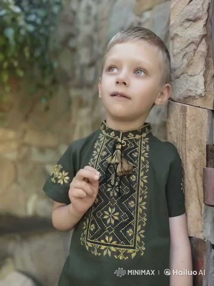 Boy wearing khaki Ukrainian embroidered shirt with short sleeves and cross-stitch design, standing against a stone wall.