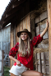 Woman wearing red Ukrainian linen blouse with traditional embroidery, standing by a wooden barn.