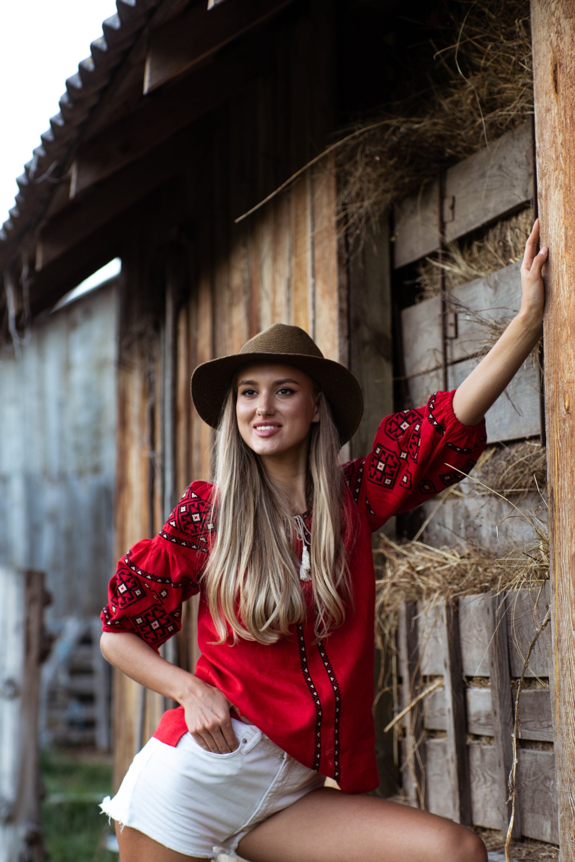 Woman wearing red Ukrainian linen blouse with traditional embroidery, standing by a wooden barn.