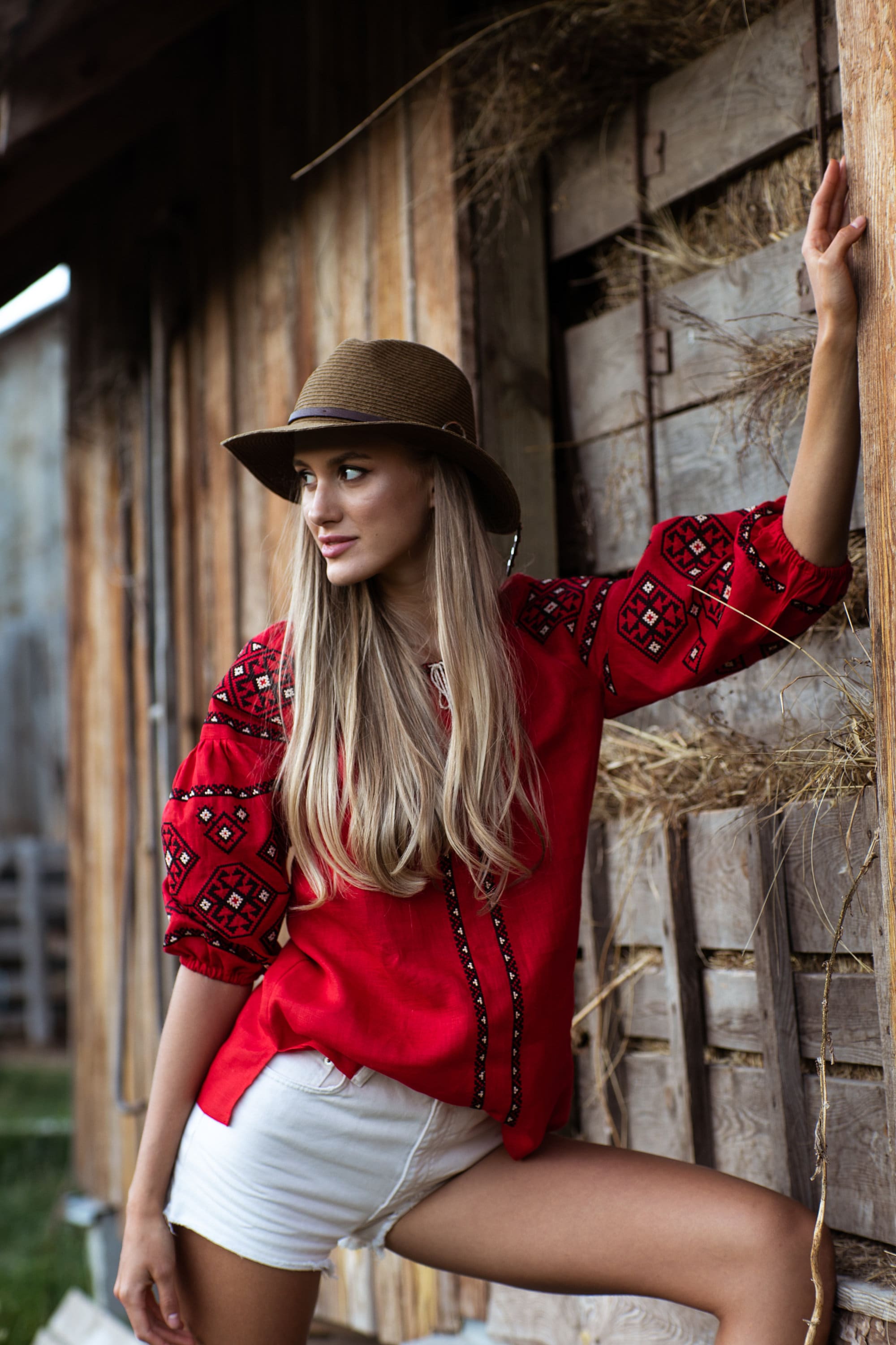 Woman wearing a red embroidered Ukrainian vyshyvanka blouse with traditional folk patterns, leaning against a rustic barn.