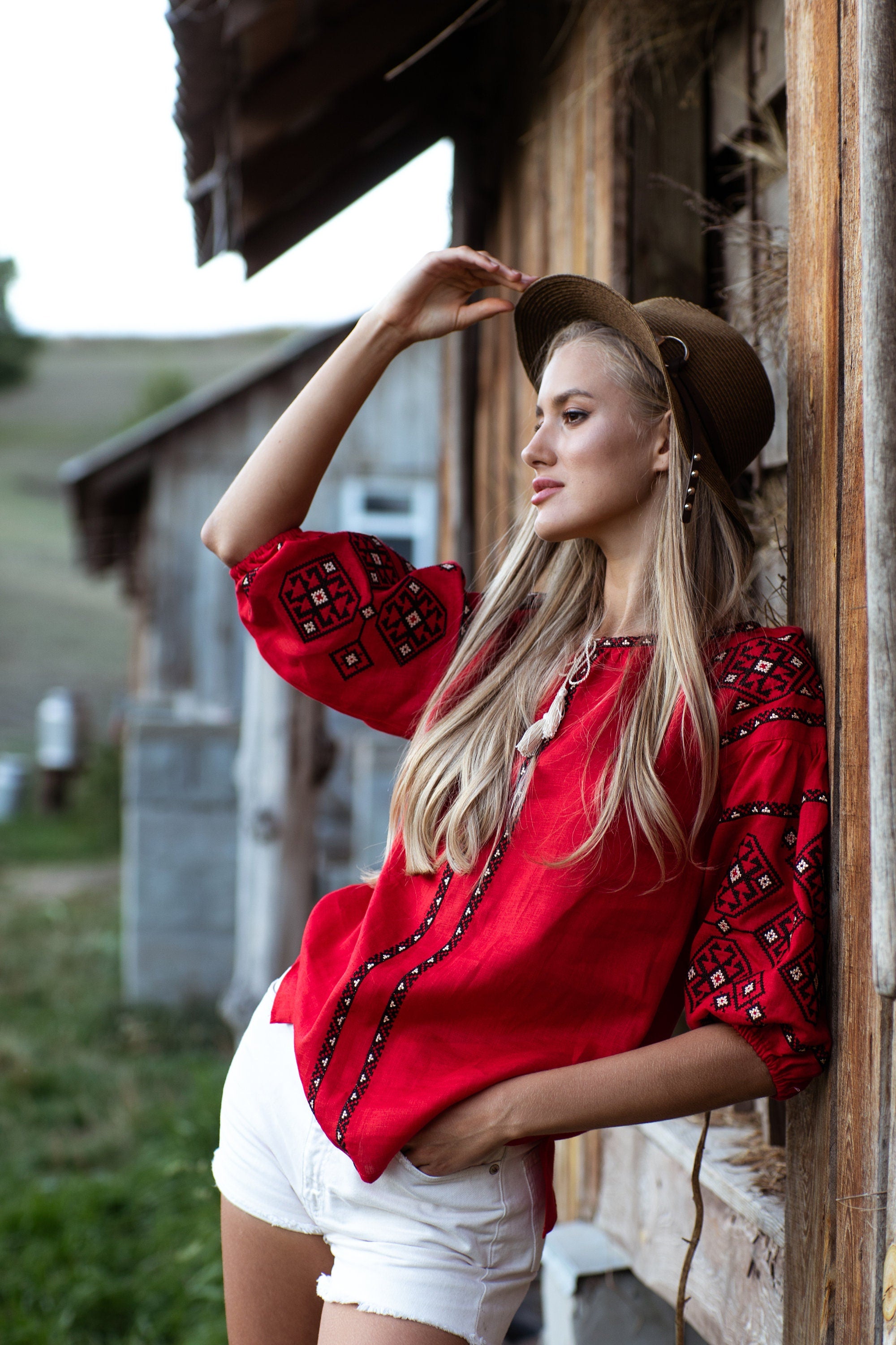 Woman wearing red embroidered Ukrainian vyshyvanka blouse, leaning against wooden building in countryside setting.