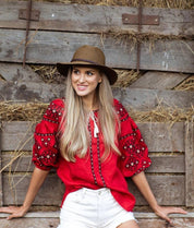 Woman wearing red Ukrainian vyshyvanka blouse with traditional embroidery, sitting outdoors against rustic wooden backdrop.
