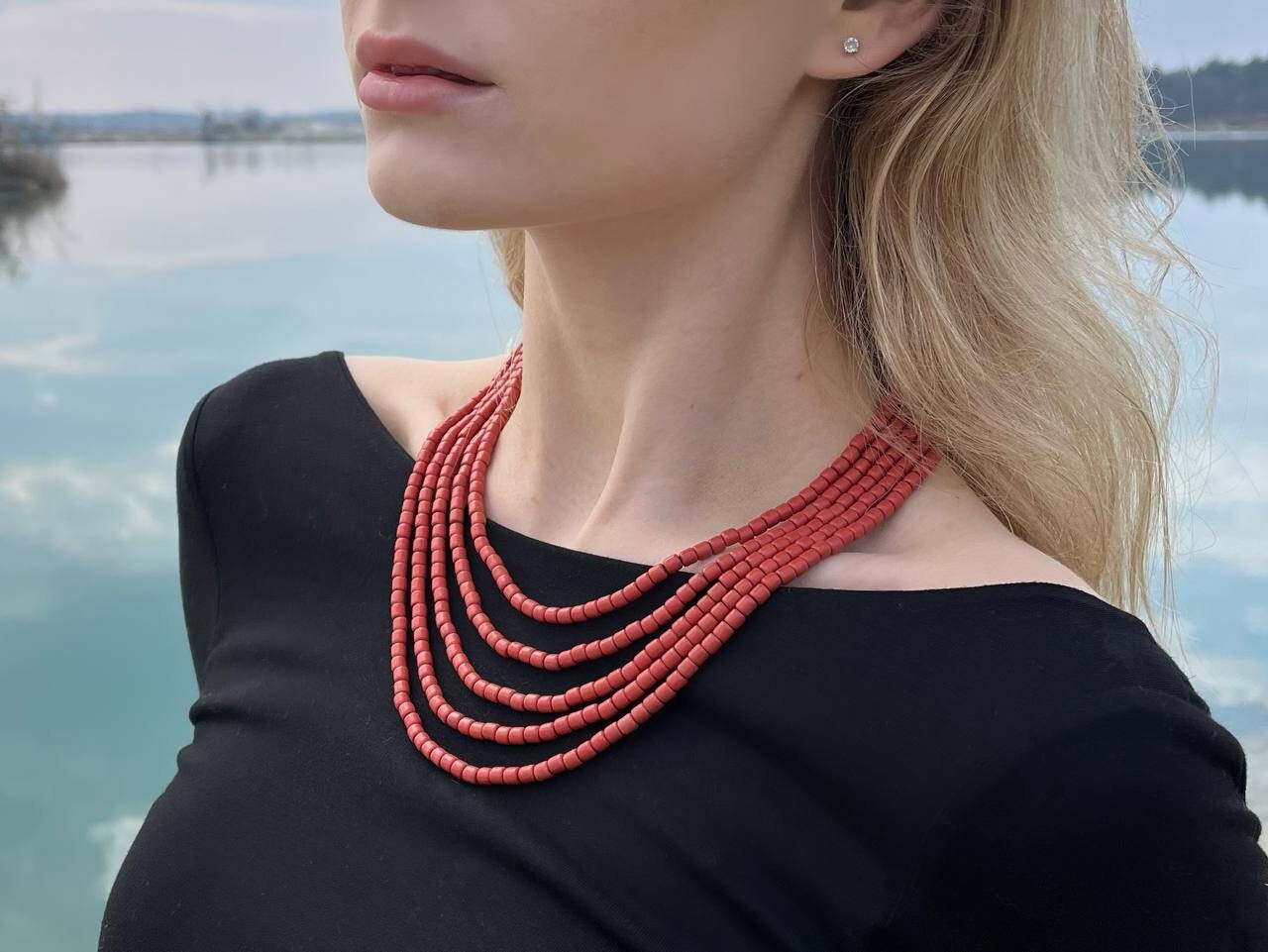 Woman wearing red ceramic beaded necklace, Scythian-inspired, on a black top against a lake backdrop, showcasing Ukrainian ethnic jewelry.