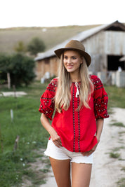 Woman in a red linen embroidered vyshyvanka blouse with Ukrainian folk design, outdoors near rustic barn.
