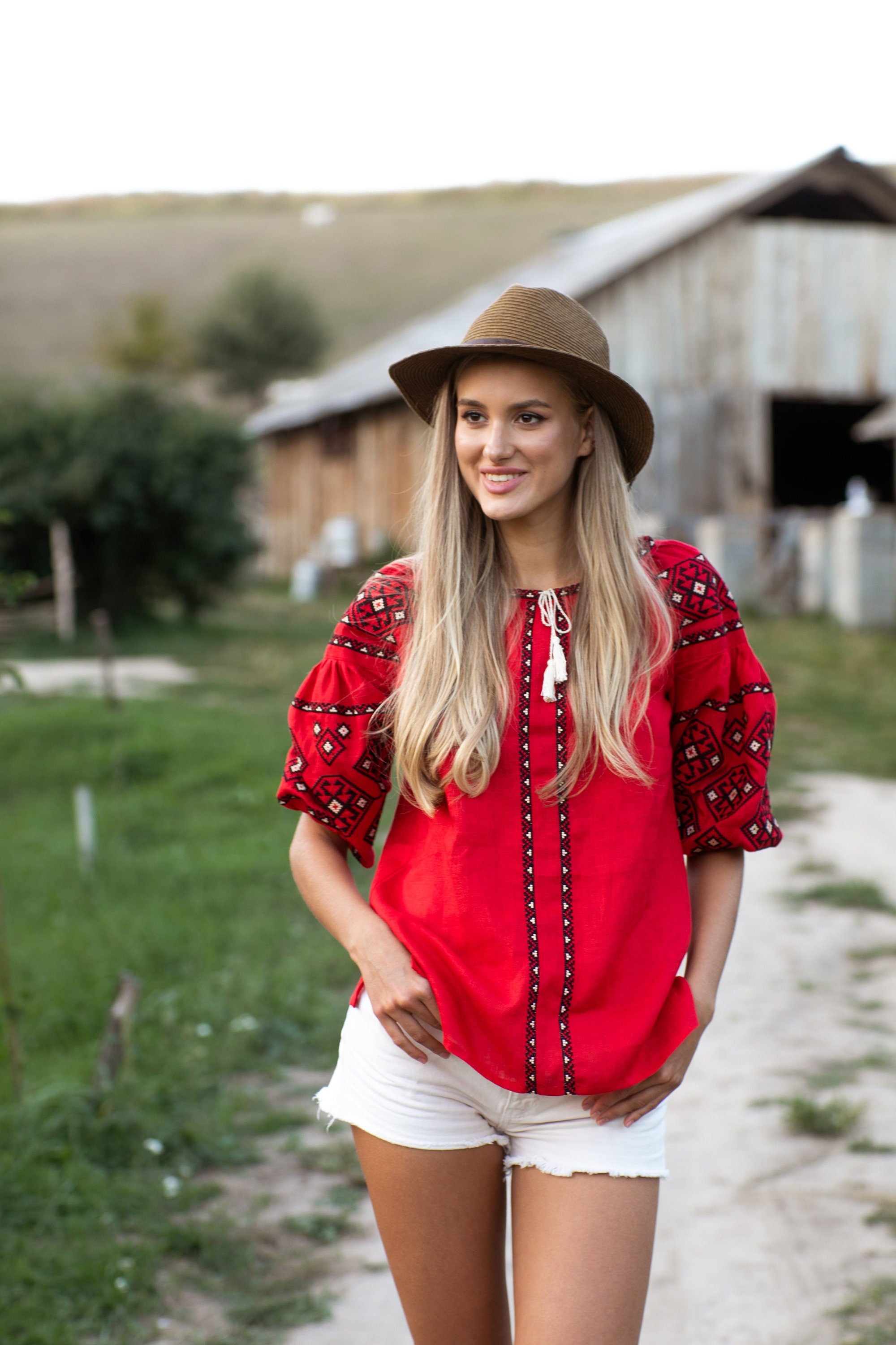 Woman in a red linen embroidered vyshyvanka blouse with Ukrainian folk design, outdoors near rustic barn.