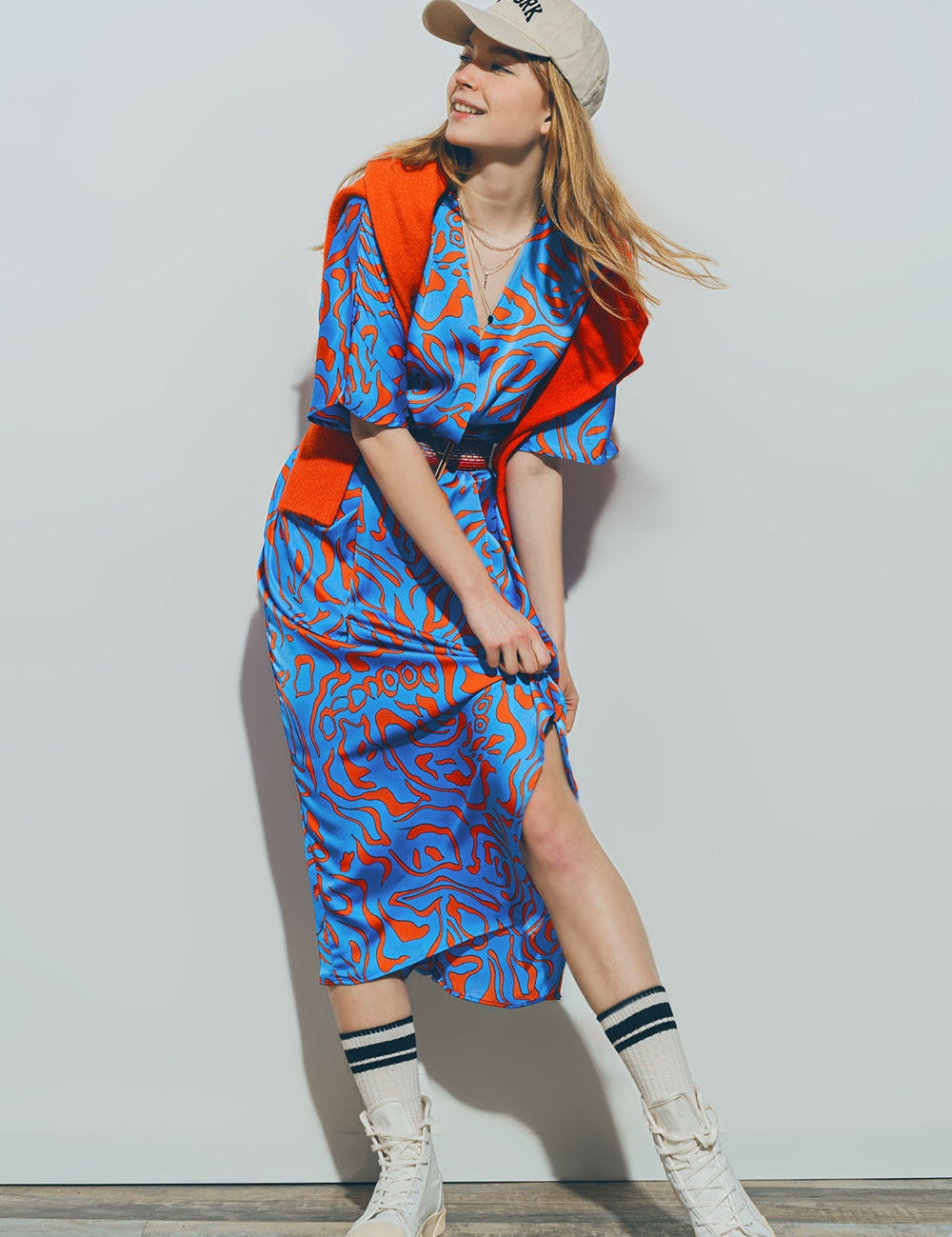 Woman in a blue and red pattern midi dress with a beige cap, standing casually with a sweater tied over shoulders.