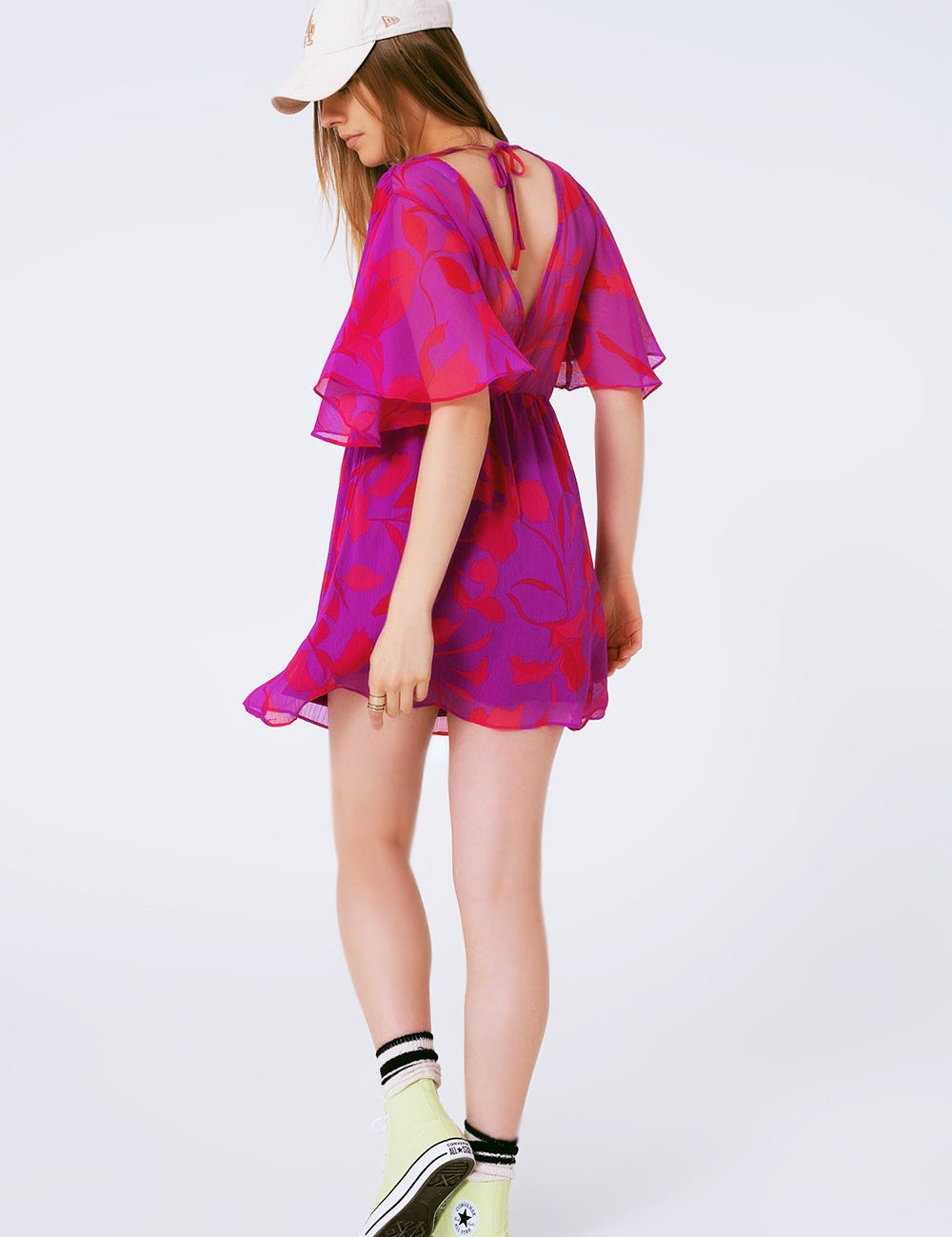 Back view of woman wearing a vibrant pink floral chiffon mini dress, paired with white sneakers and a baseball cap.