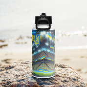 Steel water bottle with Ukrainian soccer emblem on rocky beach background, featuring vacuum insulation and wide-mouth straw.