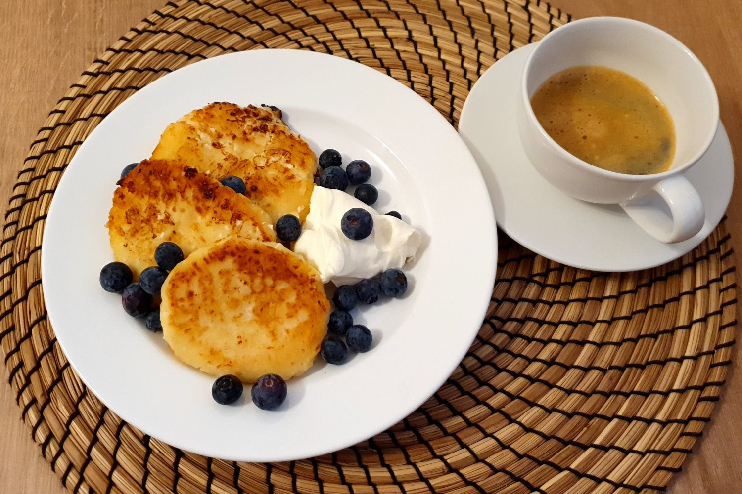 Plate of homemade Ukrainian syrnyky cheese pancakes with blueberries, sour cream, and a cup of coffee on a woven mat.