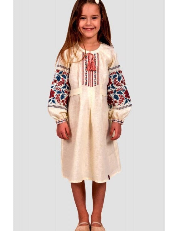 Girl in Ukrainian embroidered linen dress with floral cross-stitch, buttoned straps, and long sleeves, perfect for festive wear.